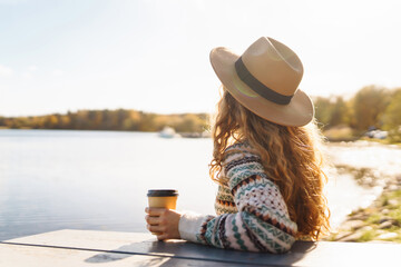 Cheerful woman in a hat relaxes, drinks tea or coffee under the sunlight against the backdrop of a lake. The concept of relaxation, freedom. - obrazy, fototapety, plakaty