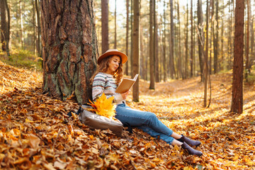 A tourist female in a hat sits in the autumn forest on yellow leaves and reads a book. Beautiful...