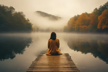 Fotobehang lonely girl sit on jetty by foggy mystic lake in autumn © krissikunterbunt