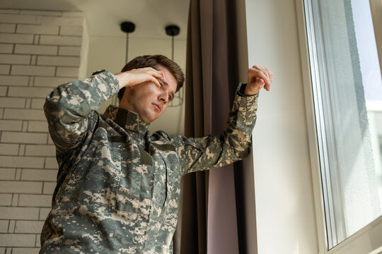 Serious Short-haired Young Man In Military Uniform Soldier Looking Through The Window, Upset Soldier Suffering From Posttraumatic Syndrome After Returning Home, Copy Space, Closeup.