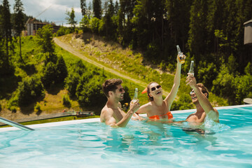 Group of cheerful friends relaxing in pool and drinking beer during vacation in mountains