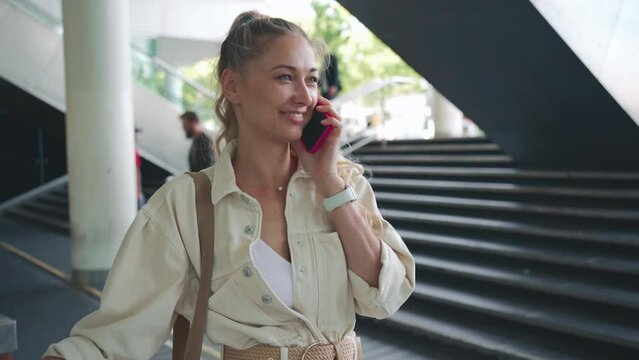 Attractive middle-aged blonde woman using cell phone in lobby business center. Cheerful woman speaking mobile phone standing in entrance hall modern building on background staircase and escalator