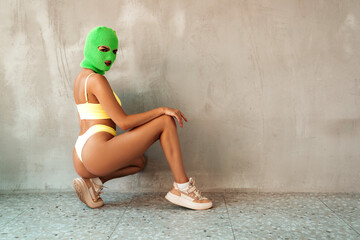 Beautiful sexy woman in green underwear. Model wearing bandit balaclava mask. Hot seductive female in nice lingerie posing near grey wall in studio. Crime and violence. Sits on the floor. Sunbathed