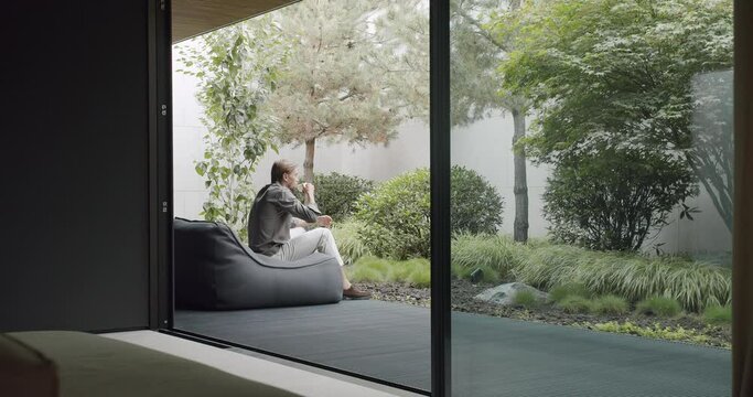 Young man sitting on comfortable bean bag chair on terrace of modern cottage house, drink coffee and enjoy morning at nature. Thoughtful male resting outdoors with cup of tea