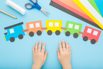 Child hands and colorful paper train with wagons on light blue table background. Pastel color....