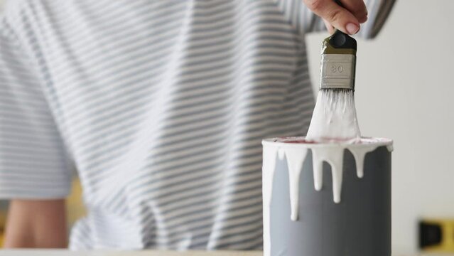 The girl paints the wooden surface of the furniture with white paint and a brush. Large drops of paint run off the brush.