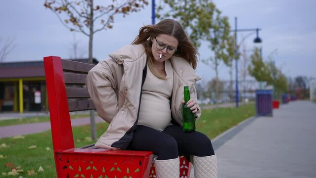 Young upset pregnant woman with beer bottle and cigarette searching lighter in pocket. Portrait of careless depressed Caucasian expectant smoking drinking outdoors on windy cloudy autumn day
