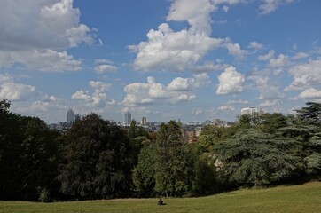 Fototapeta na wymiar Vue du parc des Buttes-Chaumont sur Paris avec ciel bleu gros nuages blancs et une personne seule assise sur l'herbe
