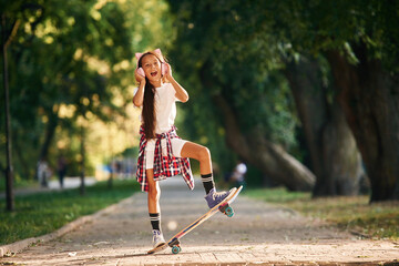 Listening to the music in cute headphones. Happy little girl with skateboard outdoors