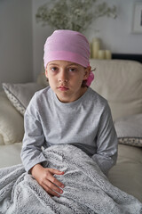 Peaceful sick little boy in bandana sitting on bed and looking a