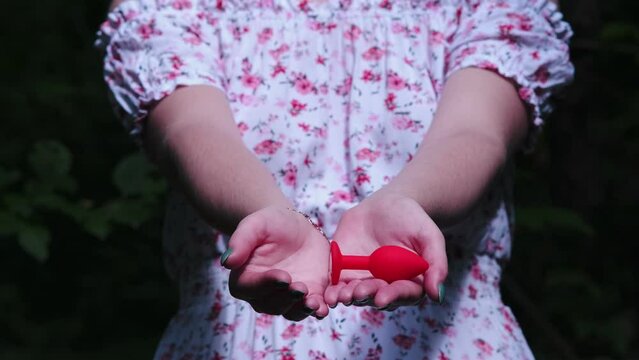 Close-up of a girl's hands in a delicate white summer dress. She opens palms, which contain a red butt plug. Sex toys, Sex shop. BDSM - outfit for adult sex games
