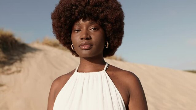 A panning-in shot captures a stunning black woman with an afro hairstyle and a white dress, gazing into the camera in the desert
