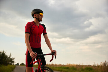 Sportive man in helmet and sportswear standing on road with bike over blue sky background. Outdoor...