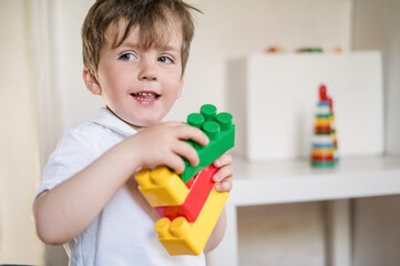 A 2-3 year old boy plays with colored plastic construction blocks sitting on the floor in a room against a white wall. child development and creativity. Logic lessons