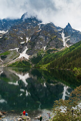 Fototapeta na wymiar A serene, cloudy mountain landscape with reflections in Morskie Oko lake and tiny tourist figures on the shore. Iconic spot in Polish Tatra National Park for nature, travel, and adventure themes