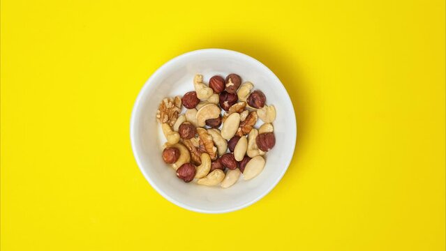 Nuts filling a bowl on a bright yellow background. Stop motion video with the cashews, hazelnuts, almonds nuts disappear from the white ceramic bowl.