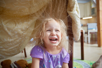 A preschooler girl laughs merrily sitting under a blanket in a shelter made at home. Electrified...