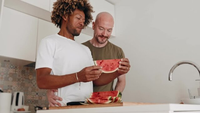 A diverse gay couple, a black man with an afro hairstyle and a bald male, share a loving moment while eating fresh watermelon in the kitchen