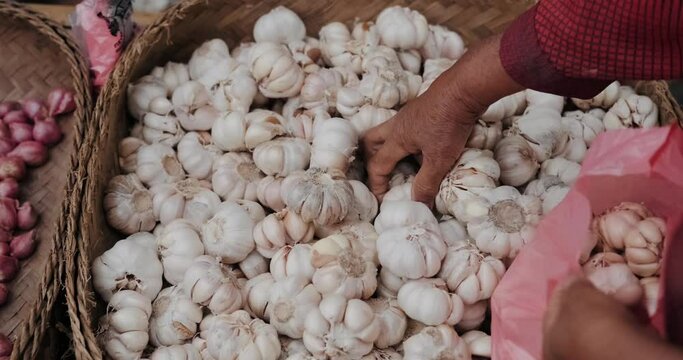 An elderly woman selects garlic from a street market and puts it in a plastic bag. Asian traditional market of organic vegetables and fruits. Buying vegetables and garlic at the market.