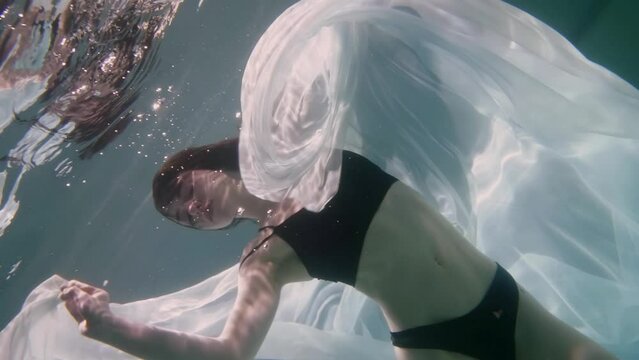 sensual young woman floating underwater, beautiful lady in black bikini, playing with white fabric