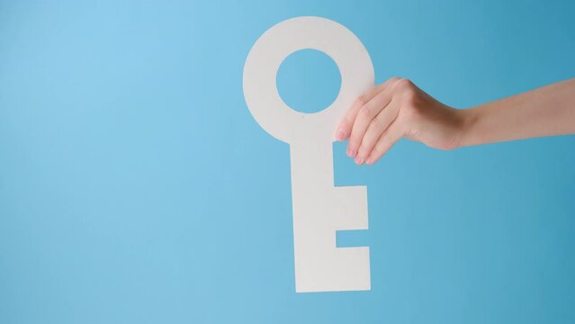 Vertical video of female hand holding big white paper key, posing over pastel blue color background wall in studio with copy space for promotion content or design. Rent, realty concept