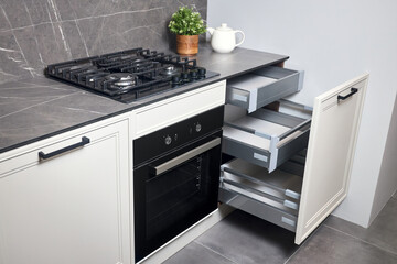Gas cooker black glass and iron hob and hood on marble light stoneware countertop with horizontal sliding pull out drawer shelves storage in kitchen cupboard.