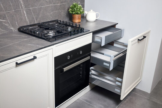 Gas Cooker Black Glass And Iron Hob And Hood On Marble Light Stoneware Countertop With Horizontal Sliding Pull Out Drawer Shelves Storage In Kitchen Cupboard.