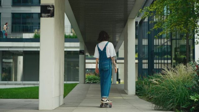 Skateboarder woman wearing blue denim overalls ride skateboard along city street, relaxed female skateboarding in downtown, urban style, modern architecture buildings, back view following slow motion