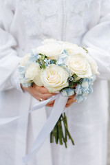 Lush fresh bouquet of blue chrysanthemums white roses, daisies with a blue ribbon in the bride's hands on the background of a wedding dress in park on a backdrop of greenery in warm summer day.