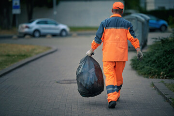 Man remove garbage from, collect garbage in plastic bag. Janitor carry black plastic bag full of garbage, refuse collector work. Worker cleanup city street, night work. Municipal worker with trash bag - obrazy, fototapety, plakaty