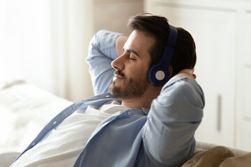 Close up calm peaceful man wearing headphones enjoying music, leaning back on cozy sofa with closed...