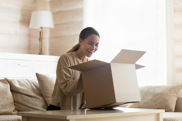 Laughing young woman looking into open cardboard box, unpacking parcel, sitting on couch in living...