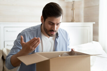 Angry irritated man unboxing parcel, looking into open cardboard box, sitting on couch at home, dissatisfied annoyed customer received wrong or broken online store order, bad delivery service concept