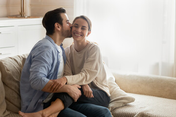Loving boyfriend kissing smiling girlfriend on cheek, enjoying tender moment, cuddling and hugging, sitting on cozy couch in living room, happy wife and husband spending romantic date at home
