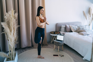 Slim woman standing and doing one legged yoga pose on mat near bed