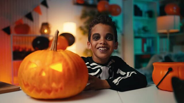 Little boy in a skeleton costume hides behind a carved Halloween pumpkin, party. Halloween holiday customs and celebration
