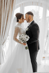 a joyful couple of young brides hug, kiss in a hotel room. The bride in a wedding dress hugs the...