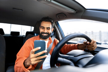 Cheerful arab driver man texting on smartphone while driving car