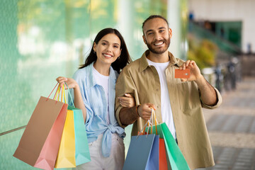 Cheerful young european guy and woman shopaholics with bags show credit card, recommend finance