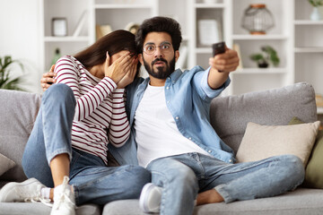 Scared millennial couple sitting on couch at home, watching horror