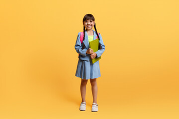 Happy asian schoolgirl with backpack and exercisebook posing on yellow