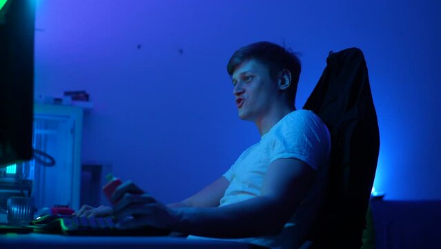 Medium shot of young man gamer inhaling of vapor with electronic cigarette. Relaxed male gamer sitting at computer monitor and playing virtual online video game with happy expression, slow motion.