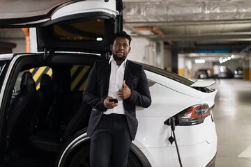 Portrait of african male in formal suit posing near car door swung up vertically with smartphone in hands, showing thumb up