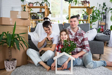 Moved family with a child and a cat pet are enjoying a new home, sitting on the sofa and holding the roof near boxes with things. Mortgage, insurance, buying and moving into your housing