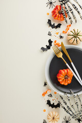 Crafting a Halloween-focused tableau for your dining experience. Top view vertical photo of plates, cutlery, skeleton hands, pumpkins, confetti, scary decor on light grey background with ad spot