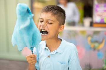 Happy boy eats blue cotton candy while visiting amusement park, spending summertime happily,...