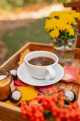 A cup of tea or coffee on a wooden tray against the background of fallen leaves, candle, autumn season, still life with leaves, flowers and chestnuts