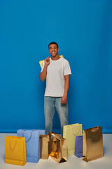 joyful african american man in casual attire holding shopping bags on blue backdrop, buying spree