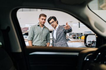 View from dark interior of car to professional auto dealer in business suit showing new vehicle to...