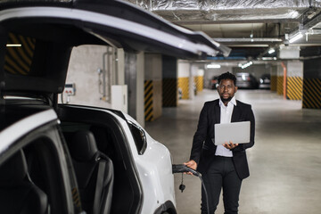 Smiling middle-aged man in formal suit plugging in EV charging cord while carrying computer in hand...